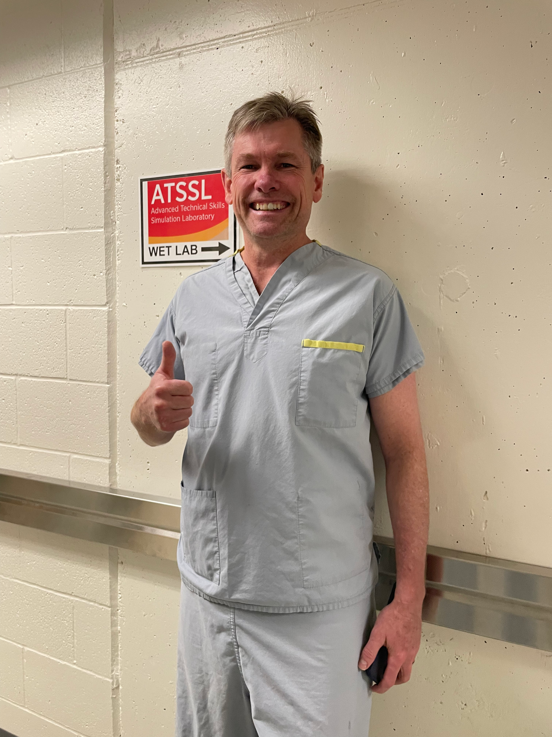 Smiling man in light gray scrubs gives a thumbs-up in a hallway near a wall sign.