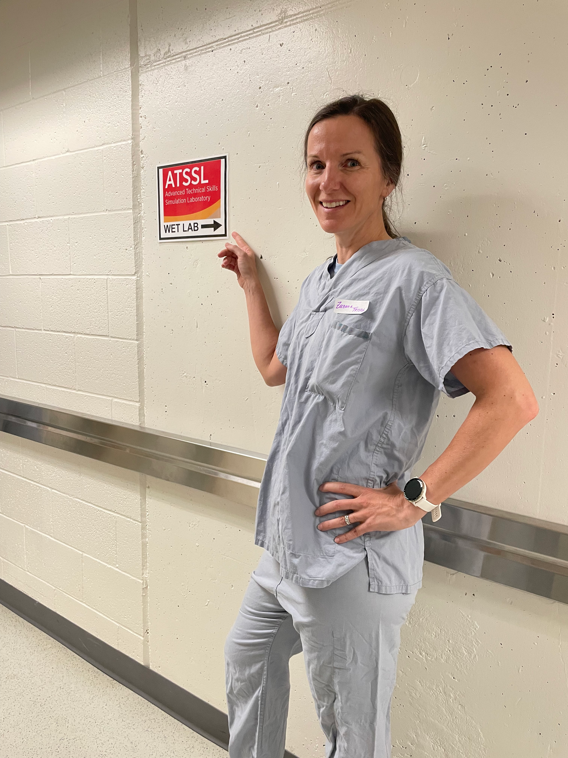 Healthcare worker in light blue scrubs pointing to a red safety sign in a hallway