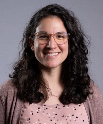 Smiling person with curly dark hair and glasses, wearing a pink patterned top and mauve cardigan against a gray background