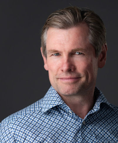 Portrait of a man in a blue patterned shirt against a dark background, looking at the camera with a slight smile