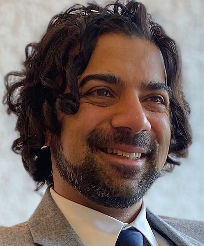 Smiling man in a gray blazer and white shirt, with curly dark hair, in a softly lit setting