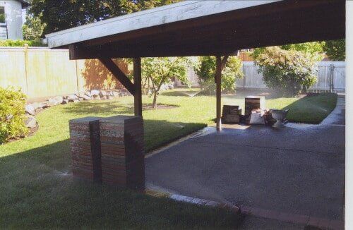 A carport with a concrete patio. Water sprays on the green grass.