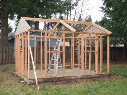 Framed wooden shed under construction in a yard, with a ladder inside.