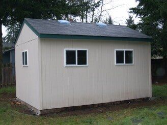 A light-colored shed with green trim and three windows sits in a grassy yard.