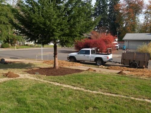White pickup truck towing a trailer with landscaping equipment near a tree in a yard.