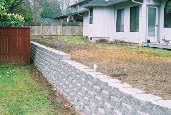 Gray retaining wall next to a house and a brown fence, separating a grassy area from a dirt patch.