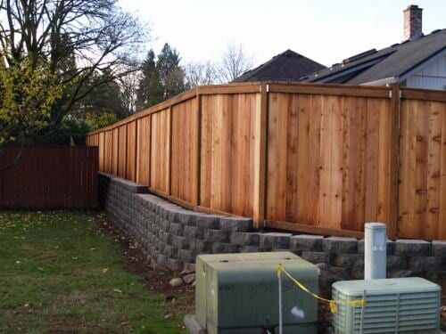 Wooden fence on a retaining wall in a backyard with utility boxes in the foreground.