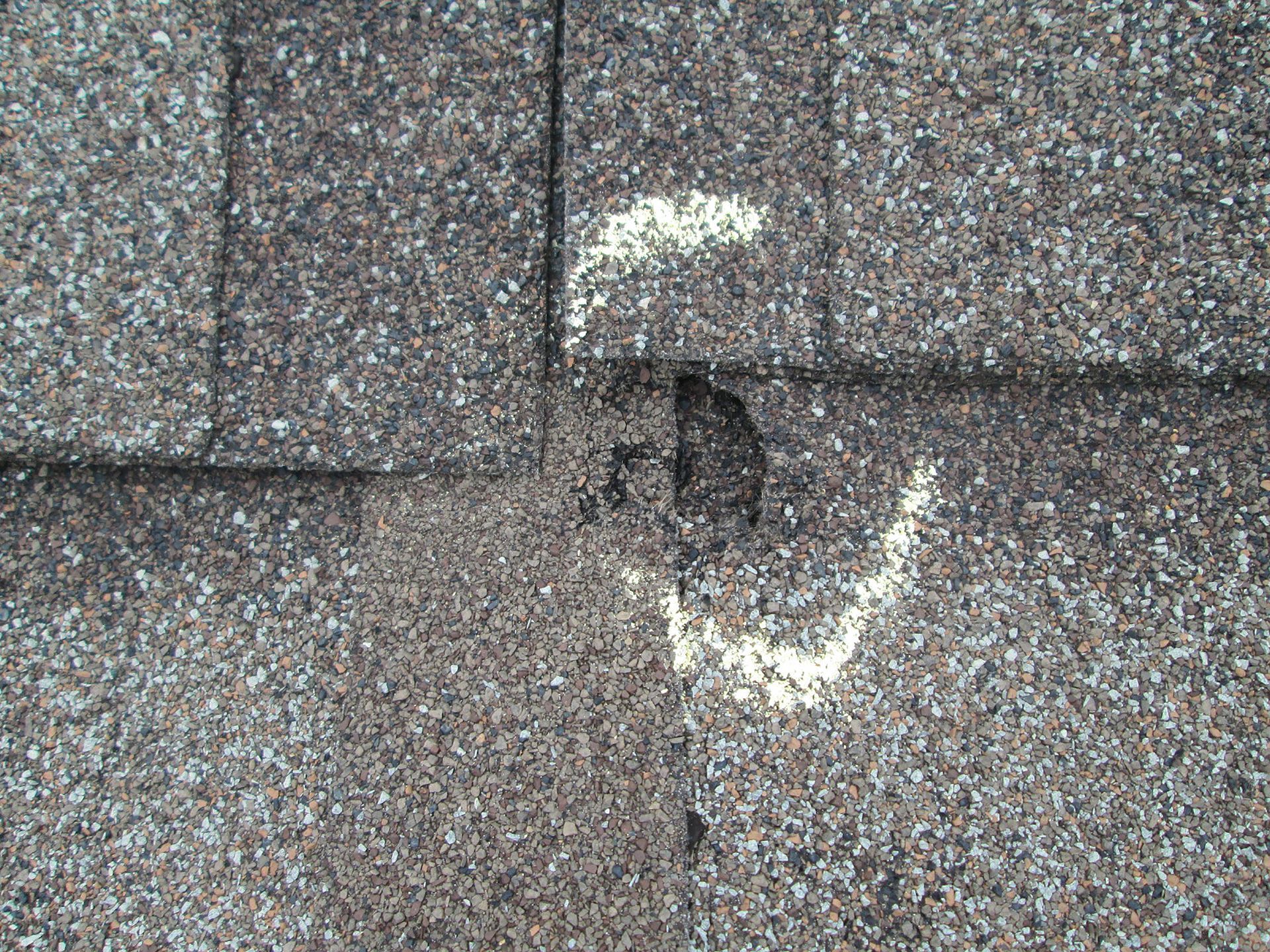 Close-up of asphalt roof shingles. White chalk marks show damage and a hole in the roof.