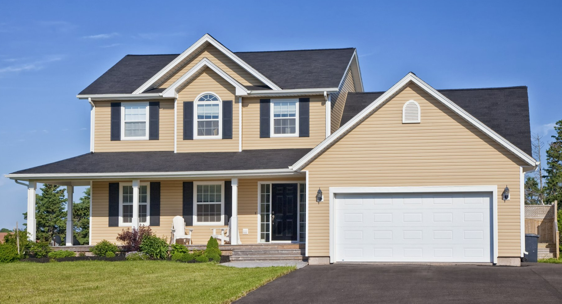 Two-story beige house with black shutters, a garage, and a porch, set against a blue sky.