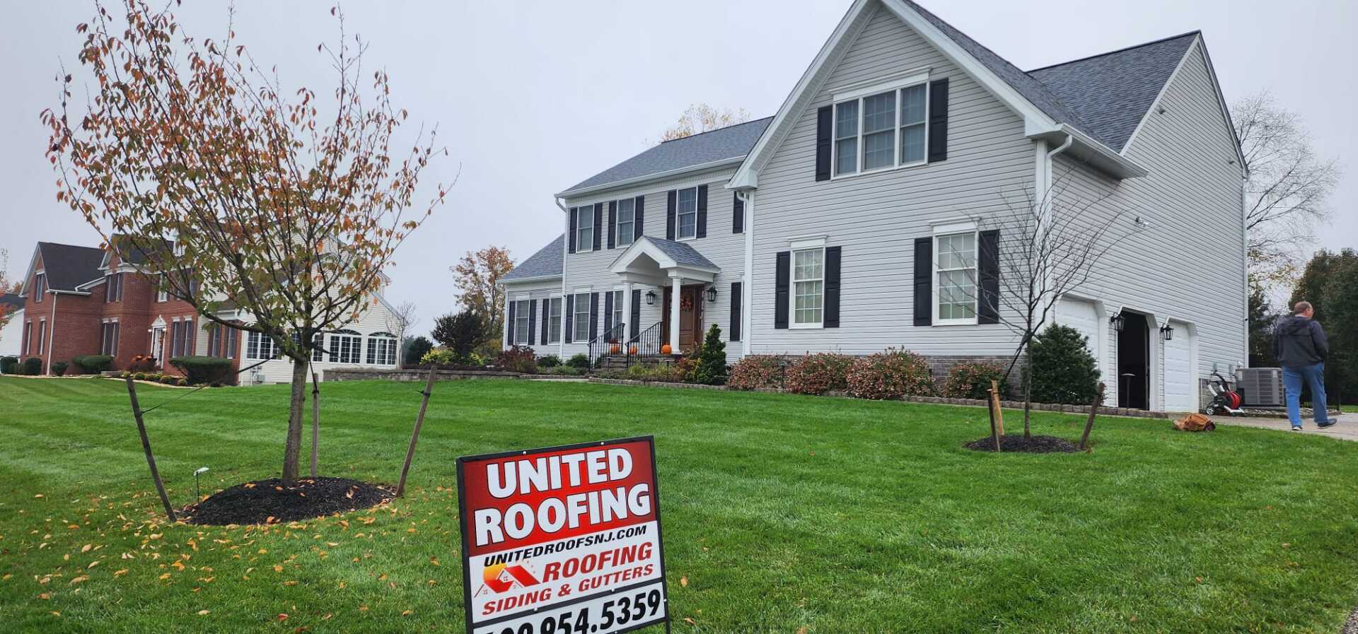 A large gray house with a United Roofing sign in the yard. Two people walk in the background. Overcast day.