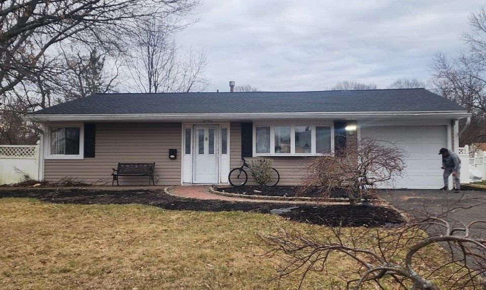 Beige ranch house with black roof, a bicycle, and a person sweeping the driveway. Overcast sky.