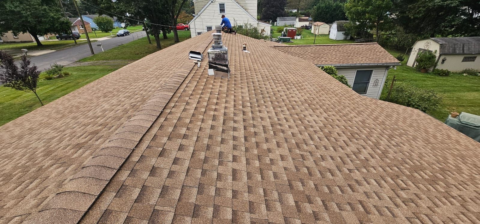 A bird's-eye view of a brown shingled roof with a chimney and surrounding trees and houses.