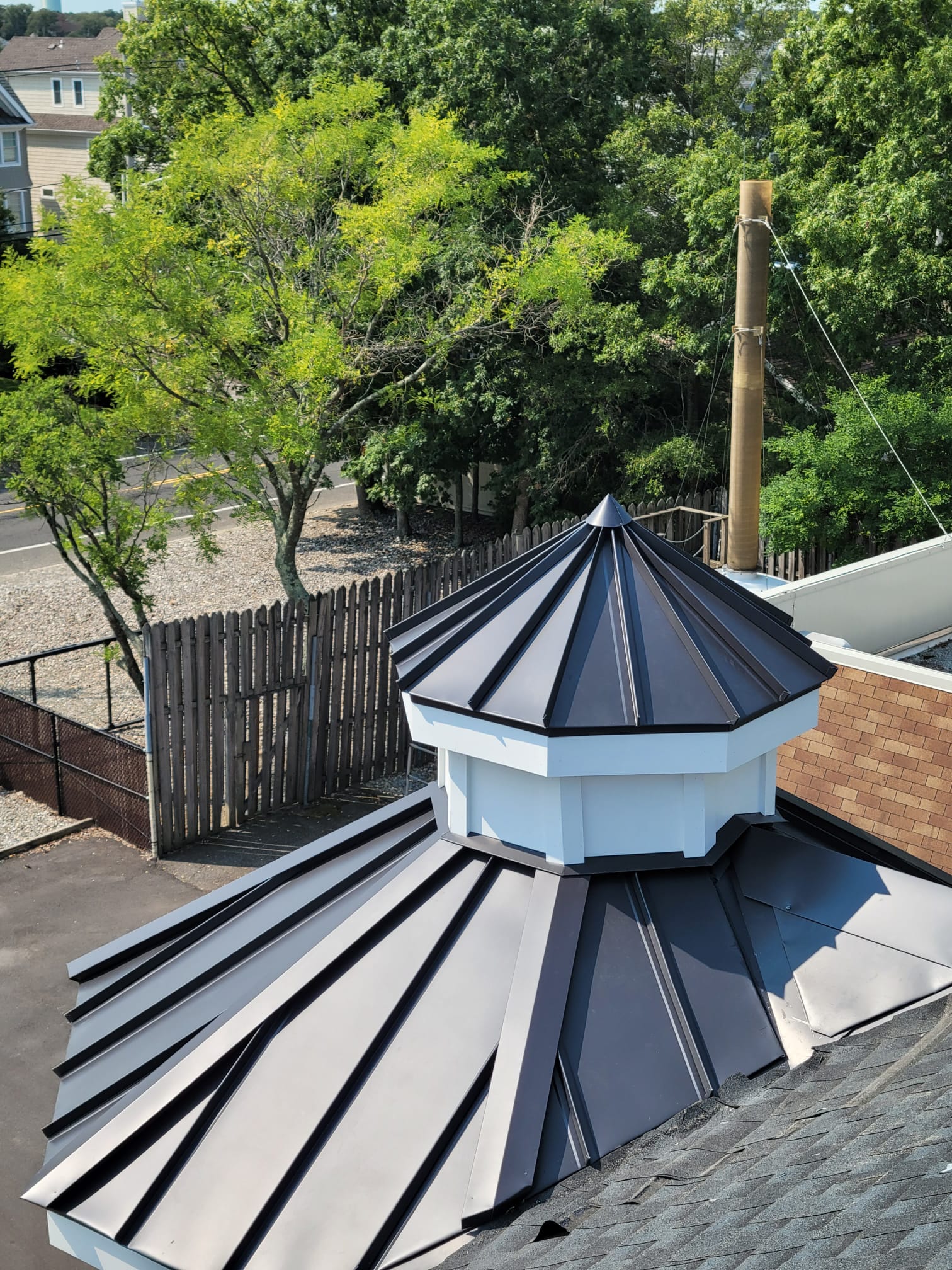 Dark gray metal roof with a white and black cupola, surrounded by trees and a wooden fence.