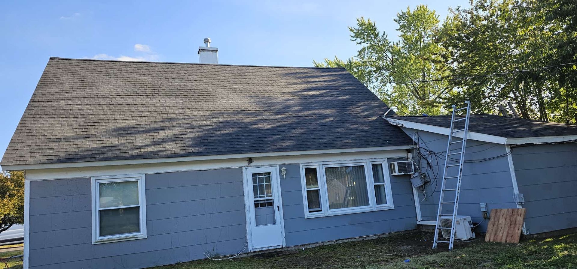 A blue house with a dark roof and a ladder propped against the side, in a grassy yard.