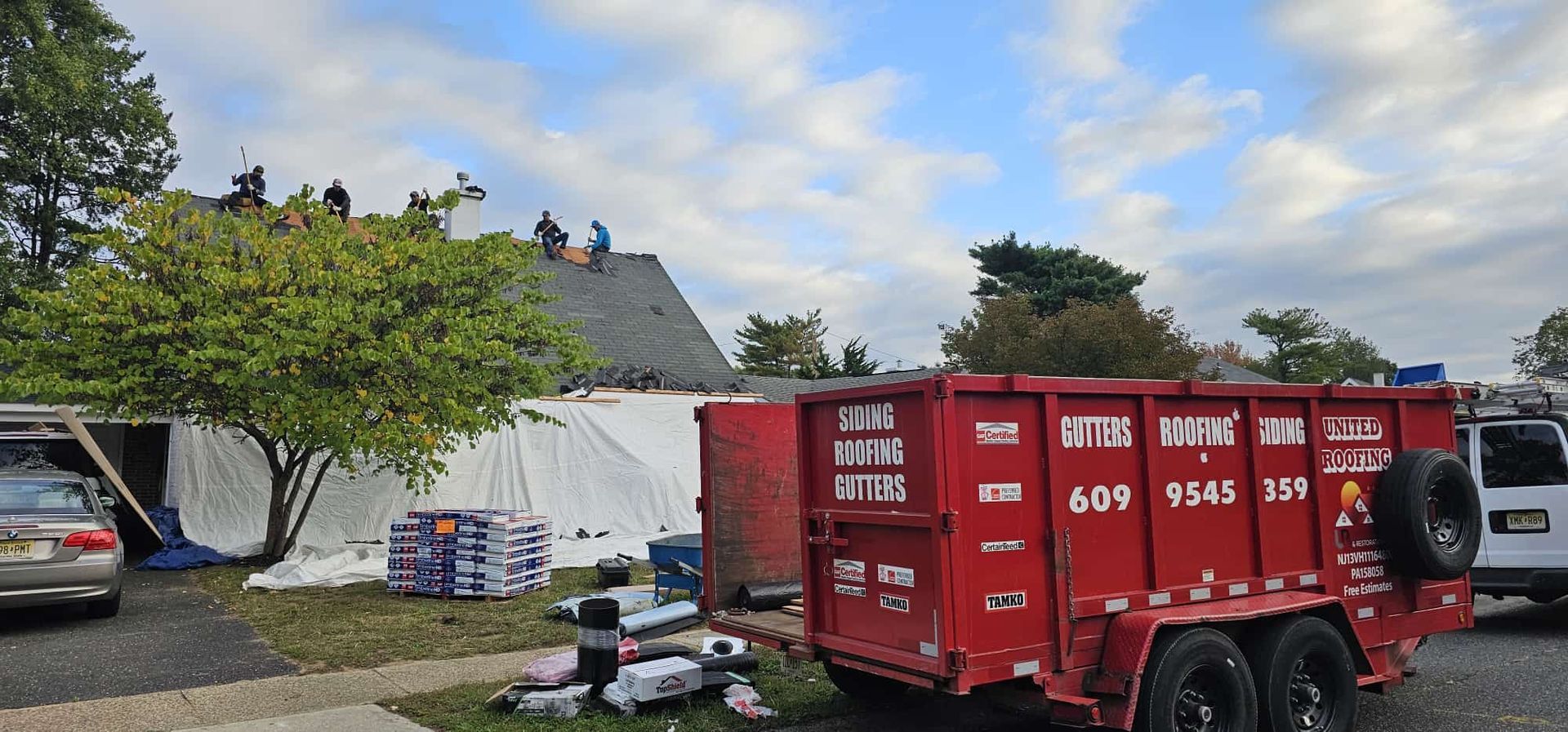 Roofers on a roof, working near a red trailer. A tree is to the left, and a car to the left of the tree.