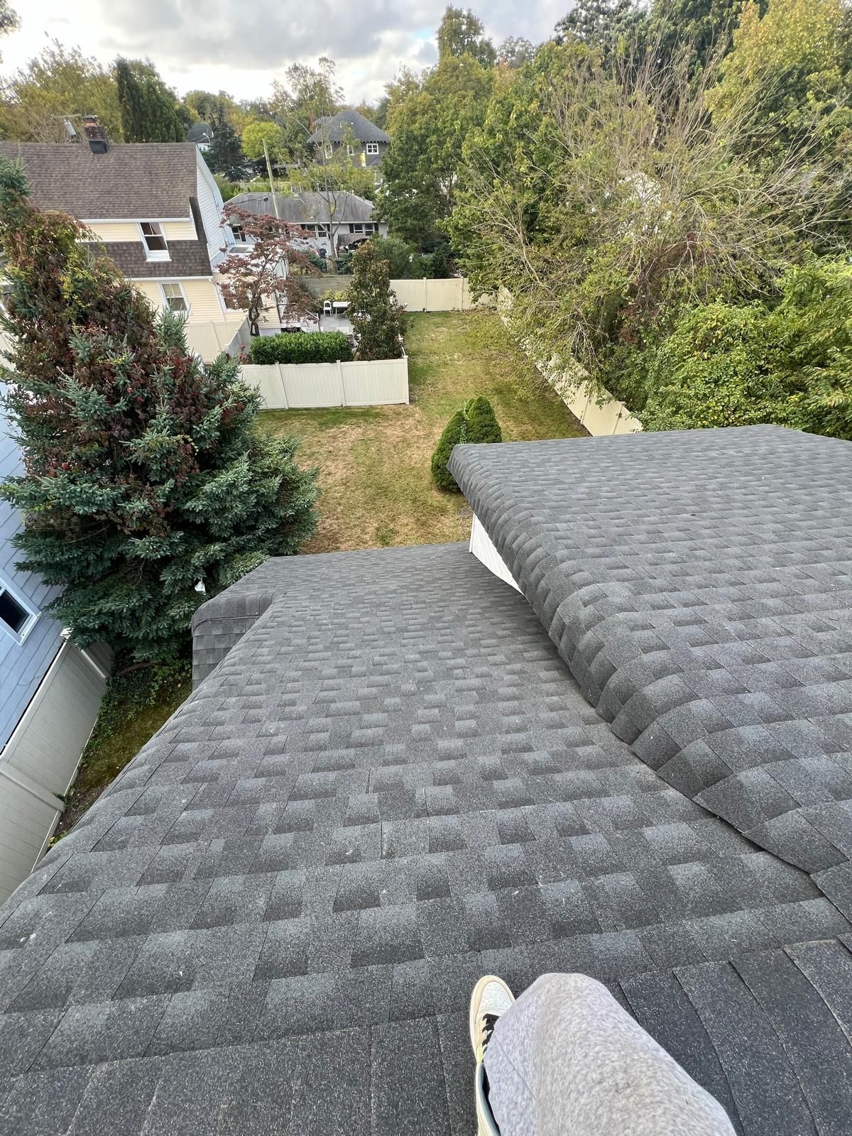 View from a rooftop: person's legs, gray shingles, yard, trees, houses, overcast sky.