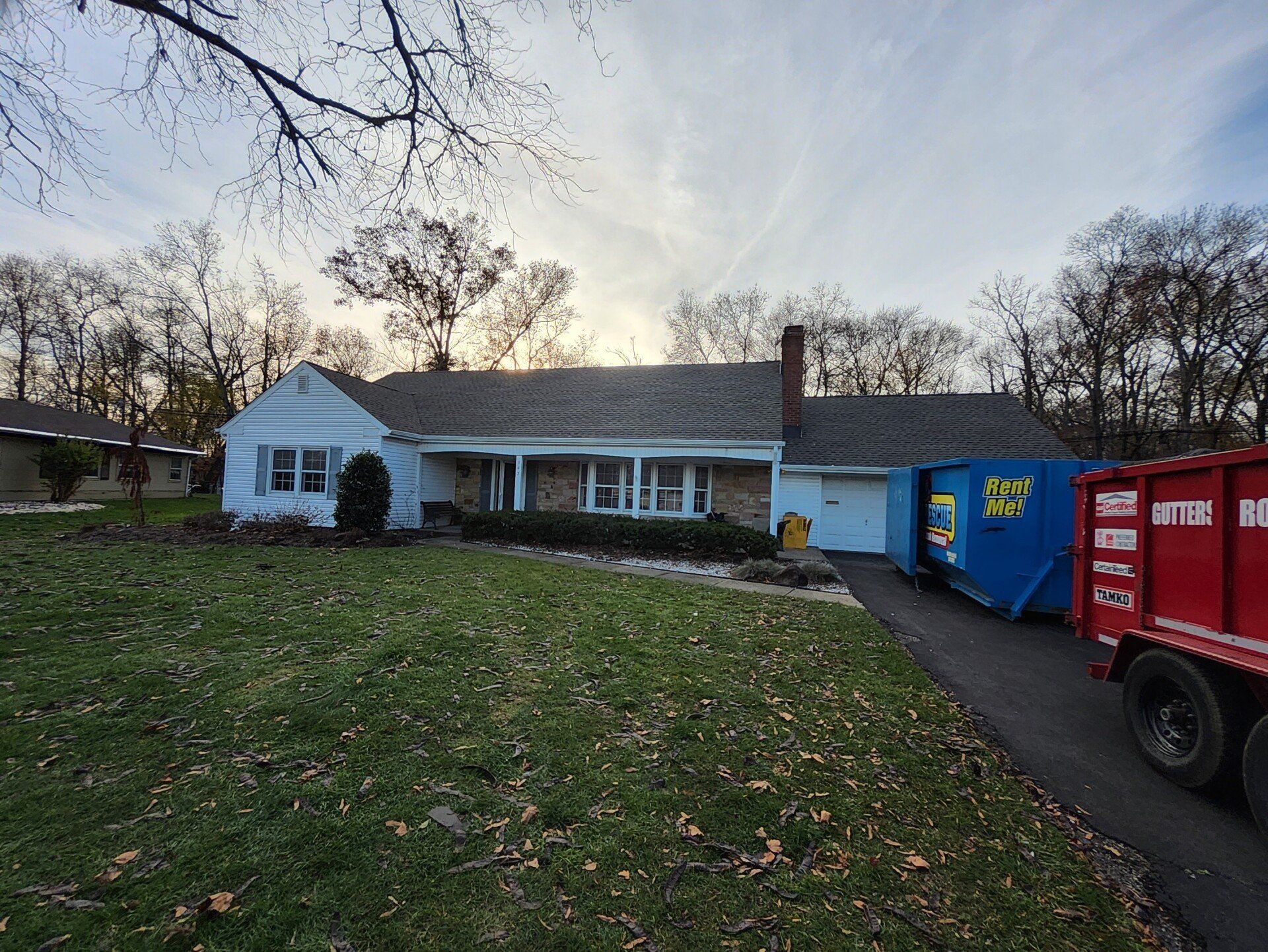 A ranch-style house with a blue dumpster in the driveway and a red truck nearby on a cloudy day.