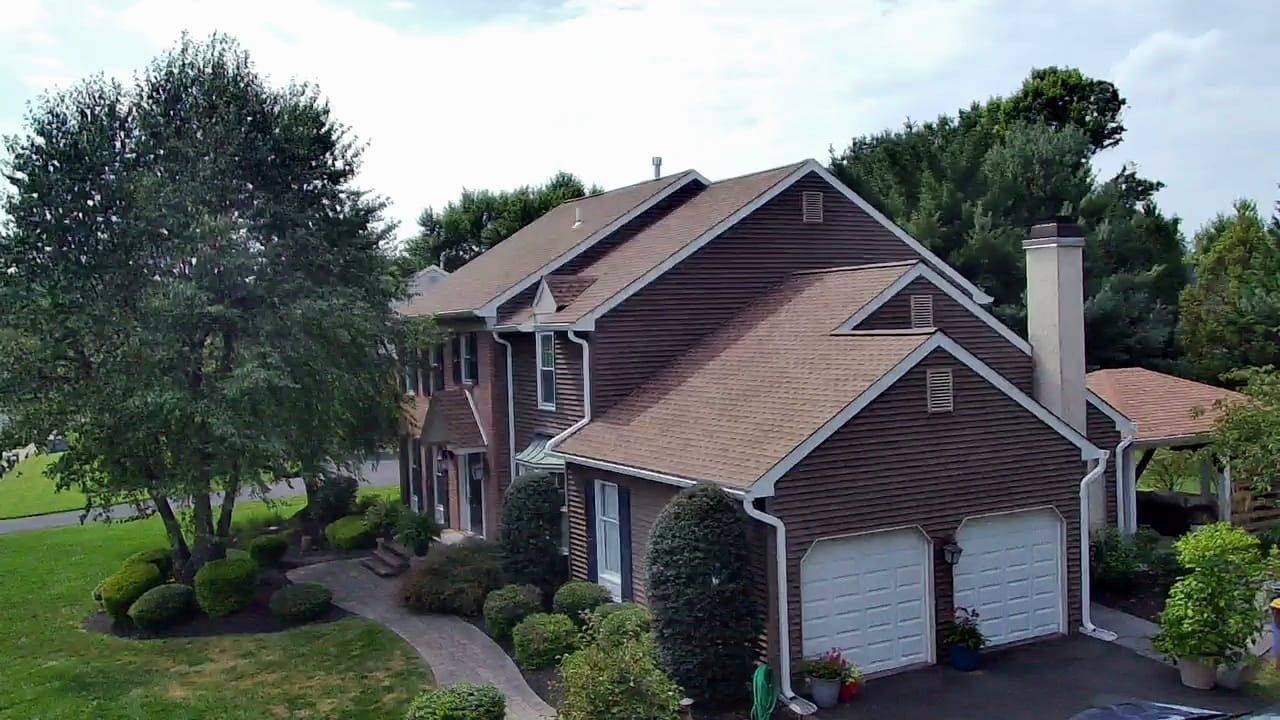 Two-story house with brown siding, brown roof, and white garage doors, surrounded by trees and green lawn.