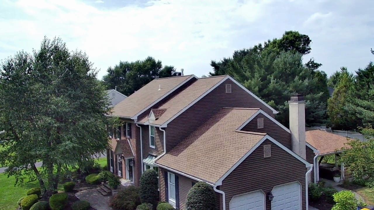 Two-story brown house with a shingled roof, trees, and a garage on a sunny day.