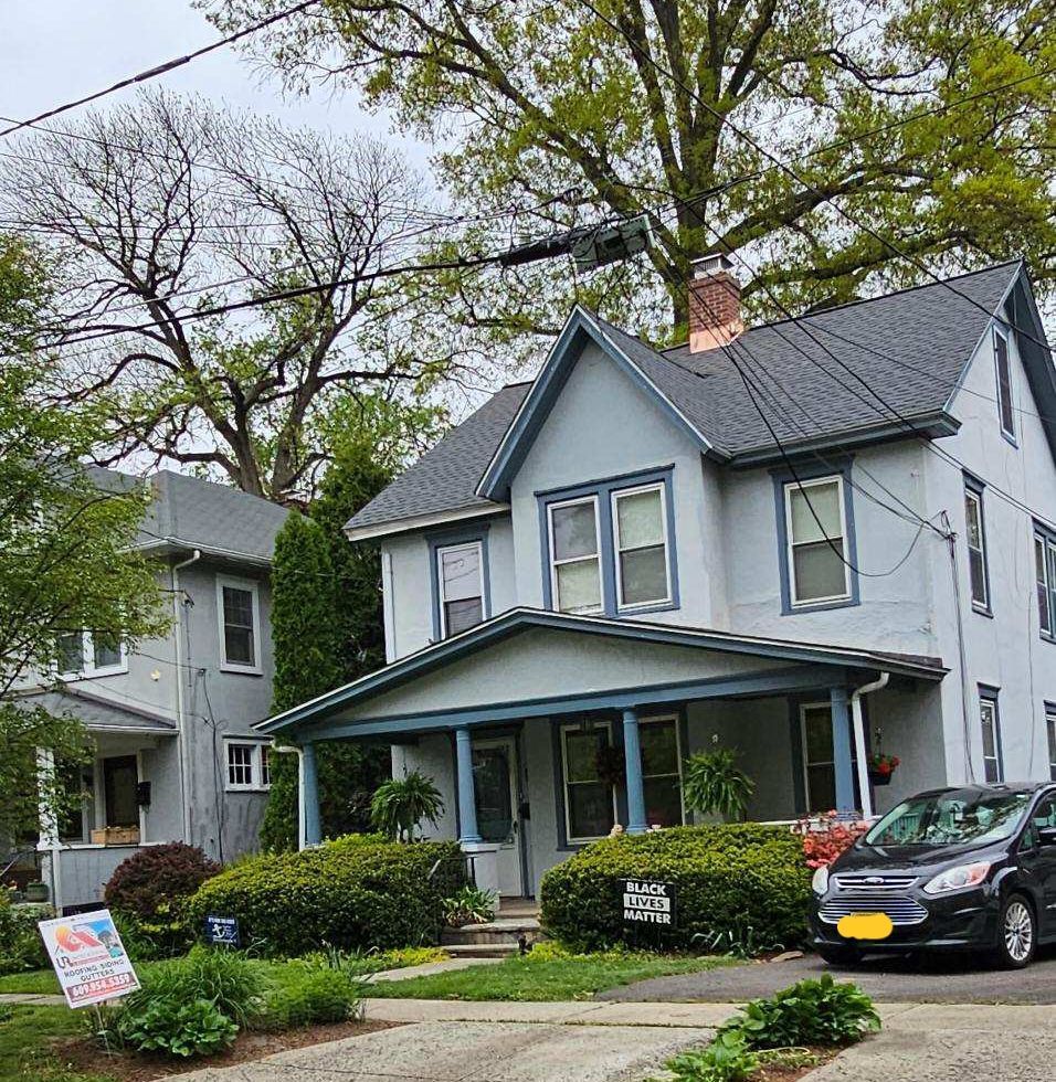 Two-story houses with blue-gray paint, front porches, and a black vehicle parked out front, on a cloudy day.