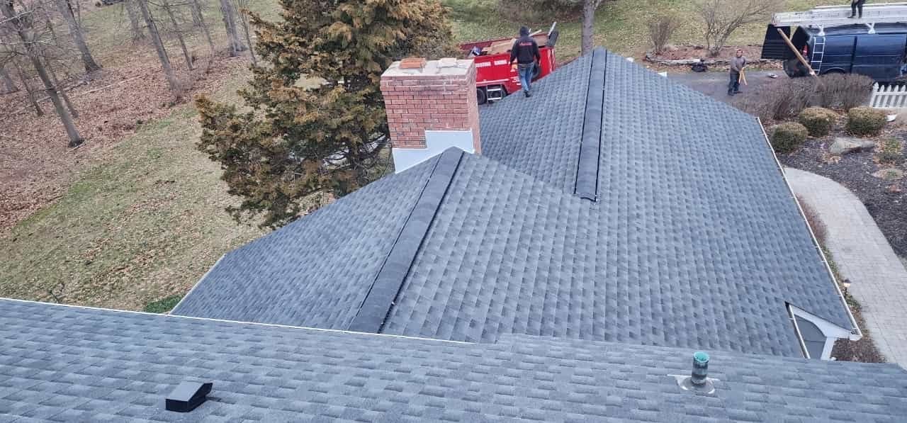 Rooftop with chimney, two people working. Red machinery, blue shingles, and trees in background.