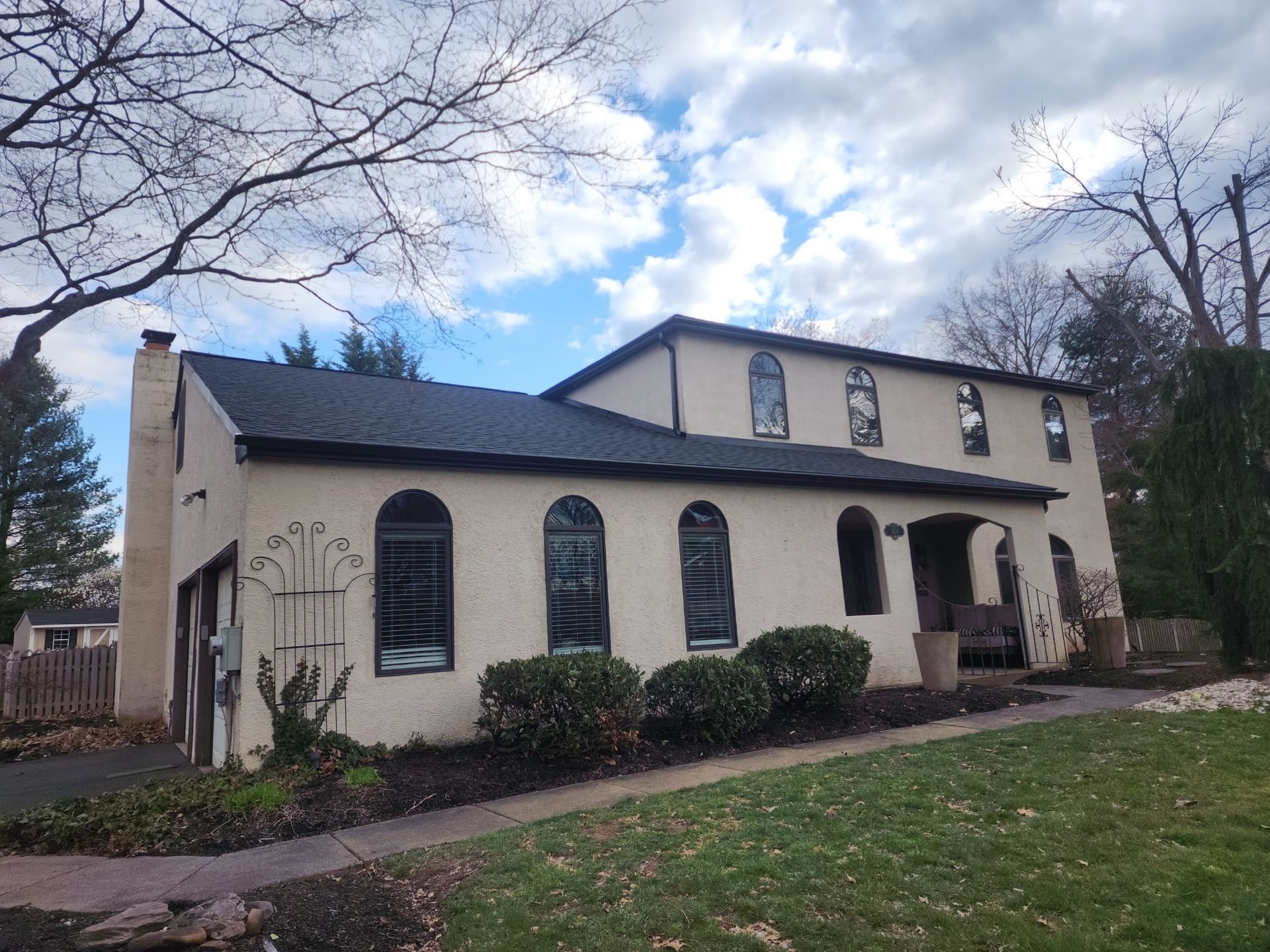 Two-story beige house with black roof, arched windows, and a grassy yard.