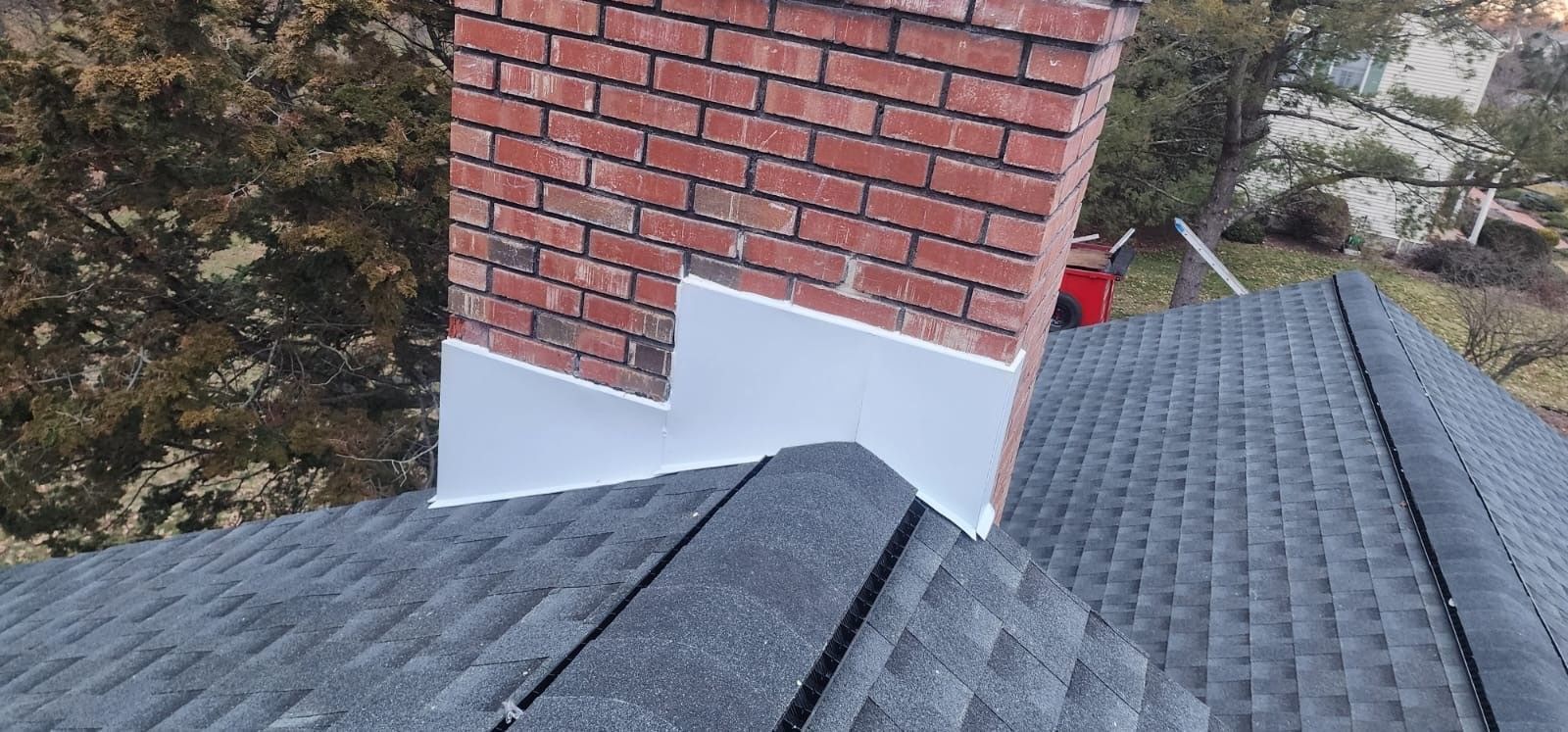 Chimney on a roof with flashing. Gray shingles, red brick, and white flashing are visible.
