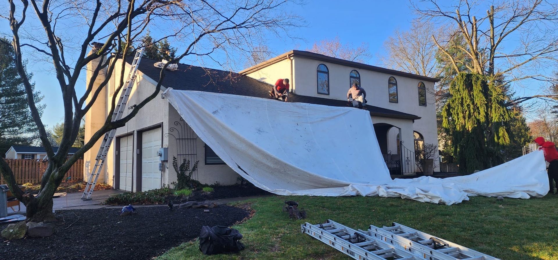 Workers on a rooftop covering a house with a white tarp. The sky is blue.