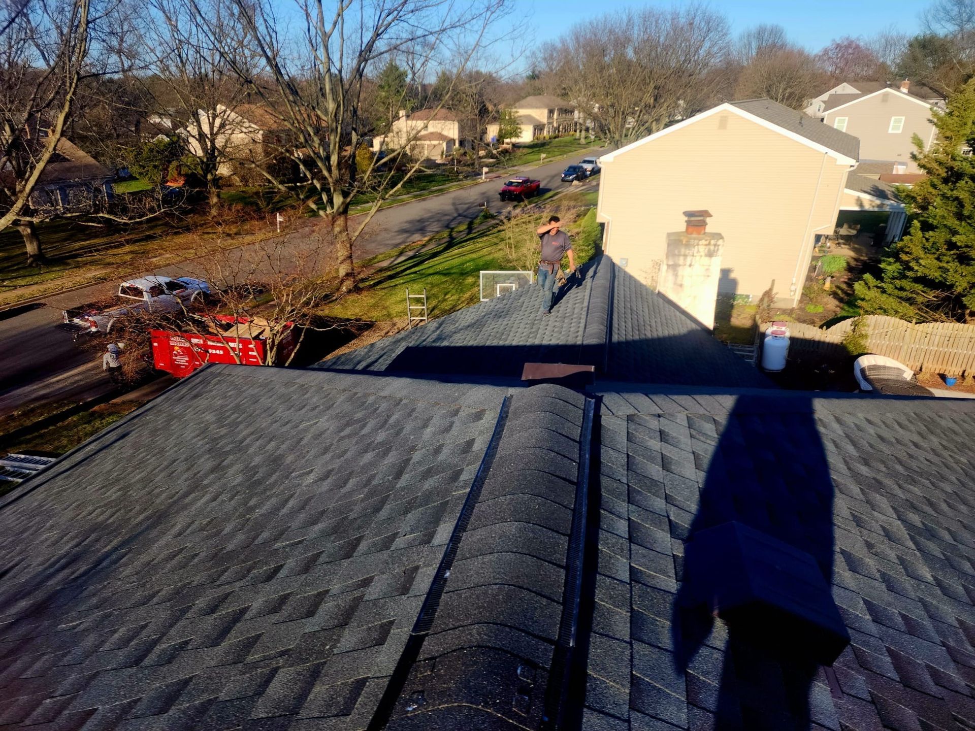 Roofer on a dark gray shingled roof with a shadow, homes, and street in the background.