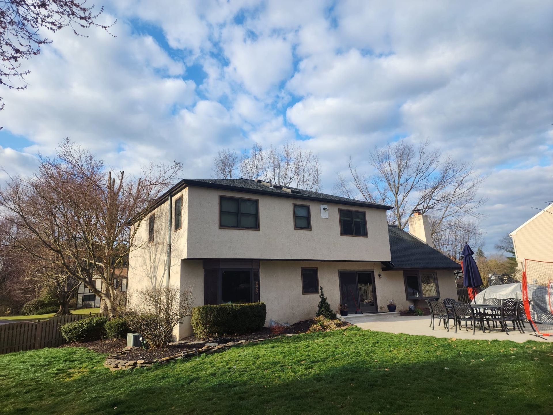 Two-story beige house with dark roof and trim, patio, and cloudy sky.