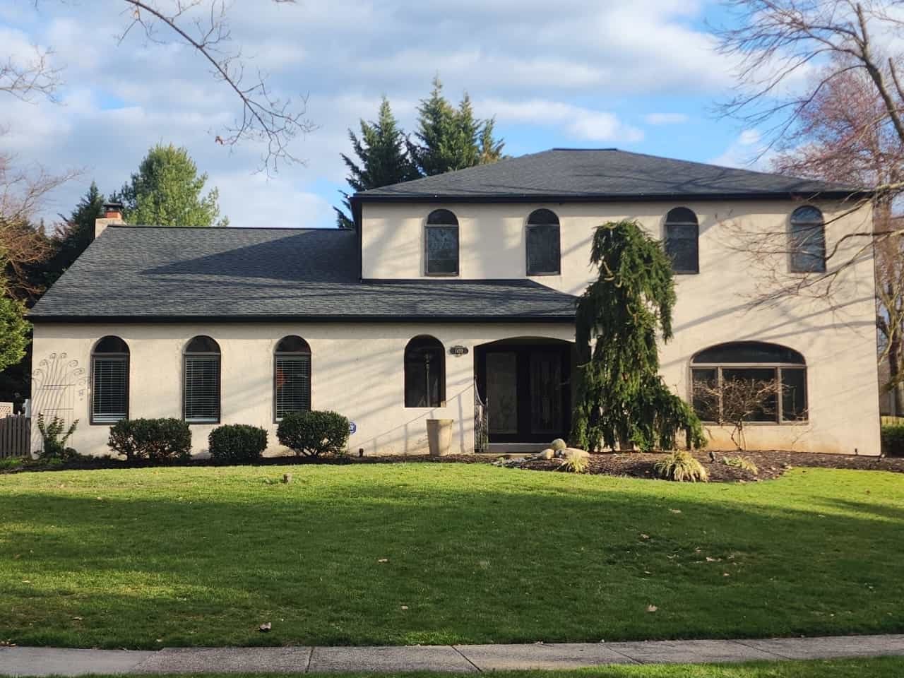Two-story beige house with dark roof and arched windows; front yard with green grass and shrubbery.