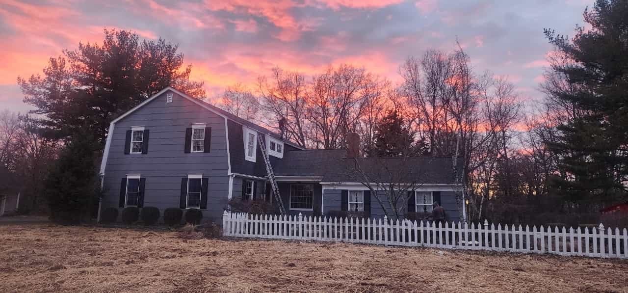 A gray house with white picket fence under a pink and orange sunset. Trees in the background.