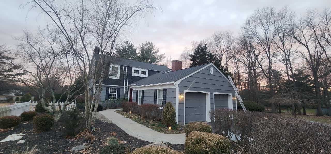 House with two-car garage, gray exterior, and a walkway, trees surrounding, overcast sky.