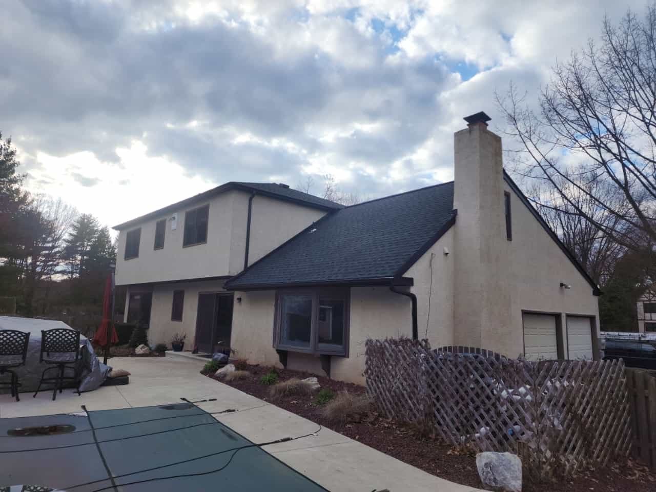 Two-story house with black roof, beige stucco siding, and a driveway next to a pool. Cloudy sky.