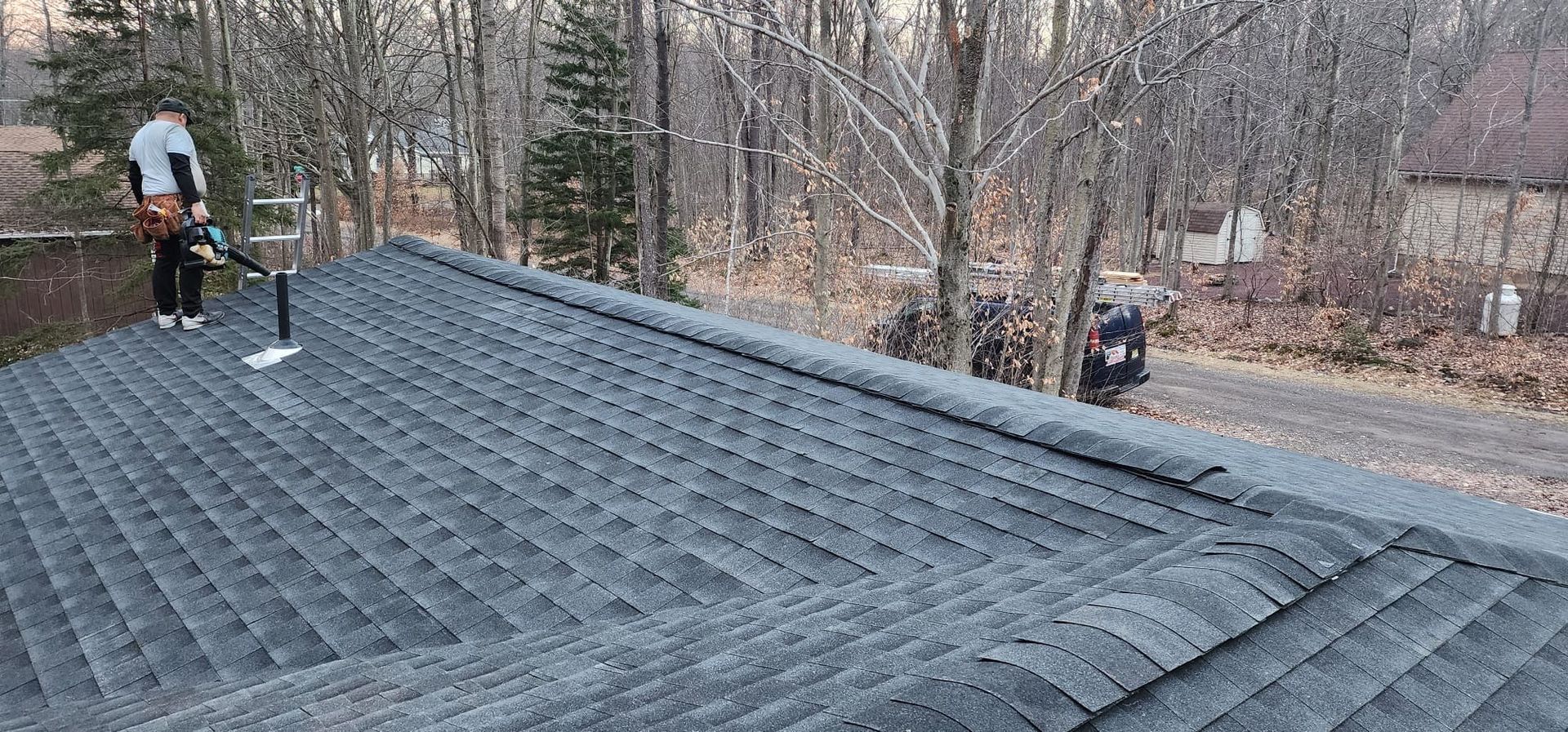 A person using a leaf blower on a dark gray shingle roof. Trees and a road are in the background.