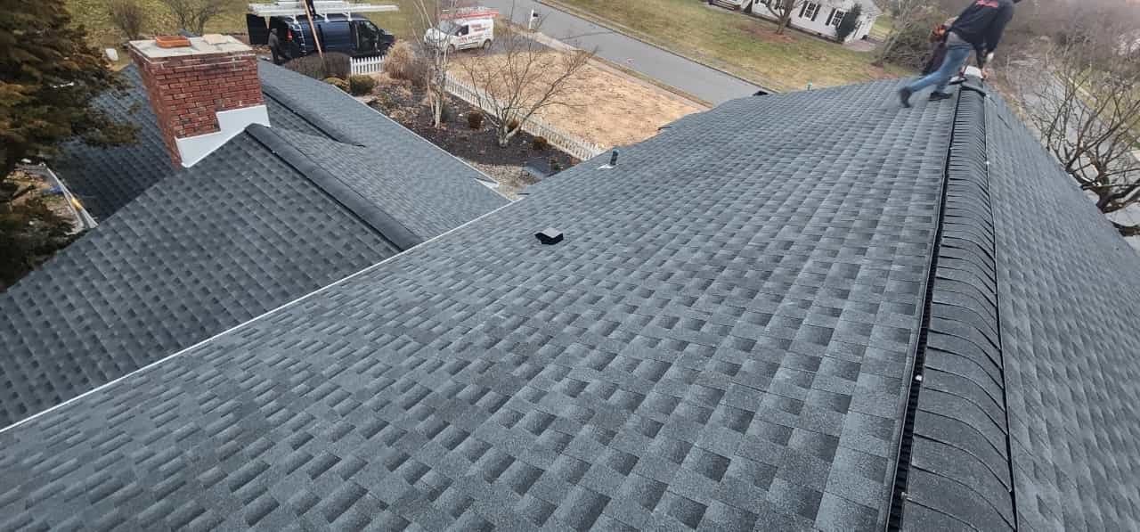 A person standing on a gray shingled roof, likely working on it. Chimney visible, street in the background.
