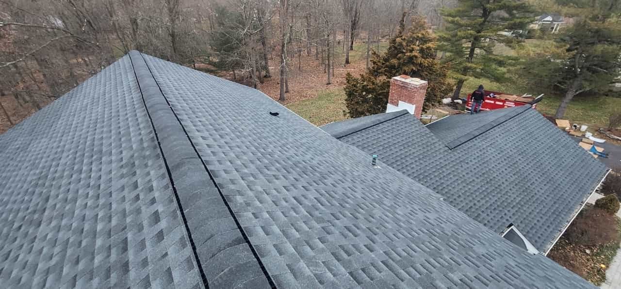 View of a gray shingled roof with a chimney and trees in the background.