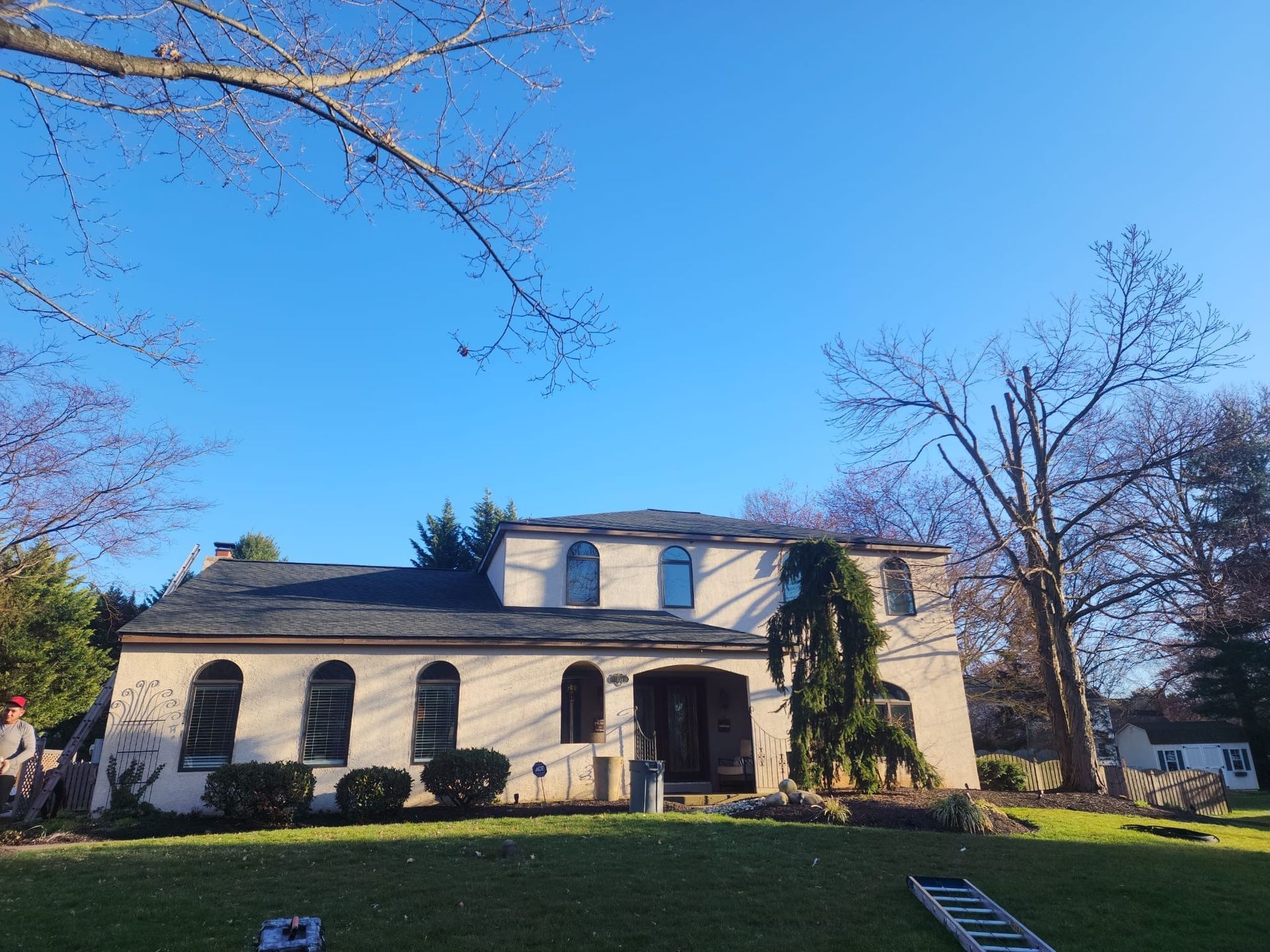 Two-story house with dark roof and light-colored walls under a blue sky, with some leafless trees and green grass in front.