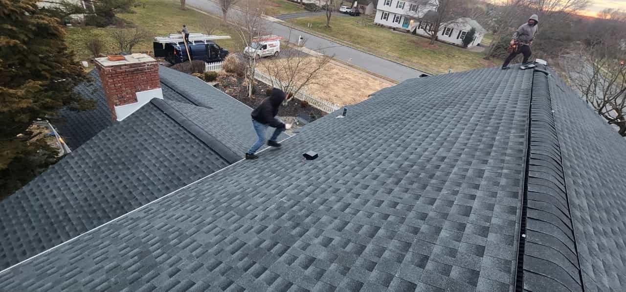 Two people working on a dark gray shingled roof, trees and houses in the background.