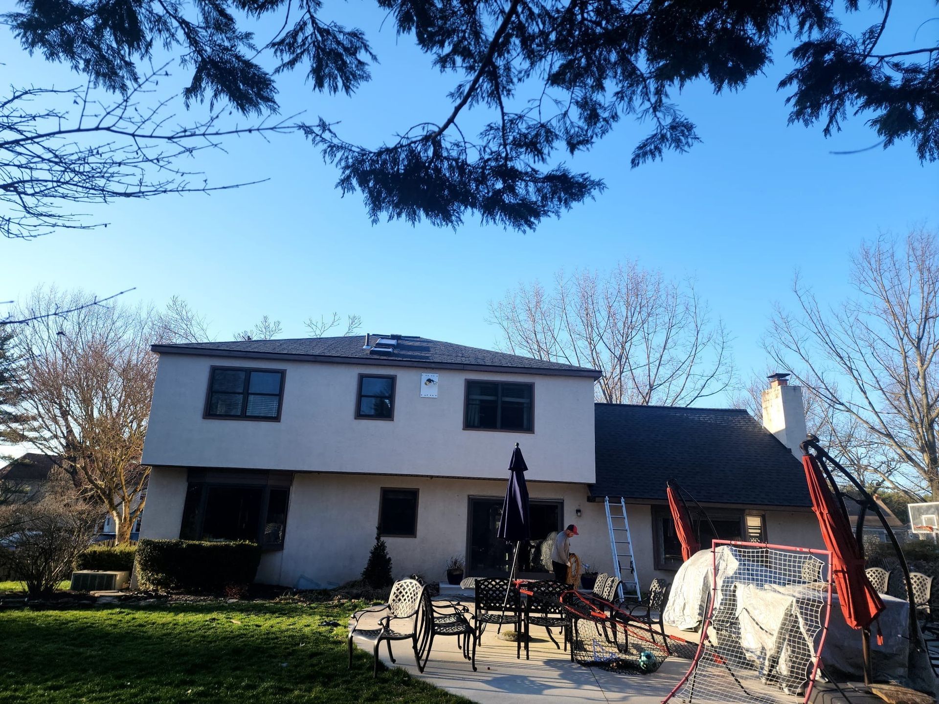 Back of a two-story beige house on a sunny day. A person on a patio near furniture. Bare trees and blue sky.