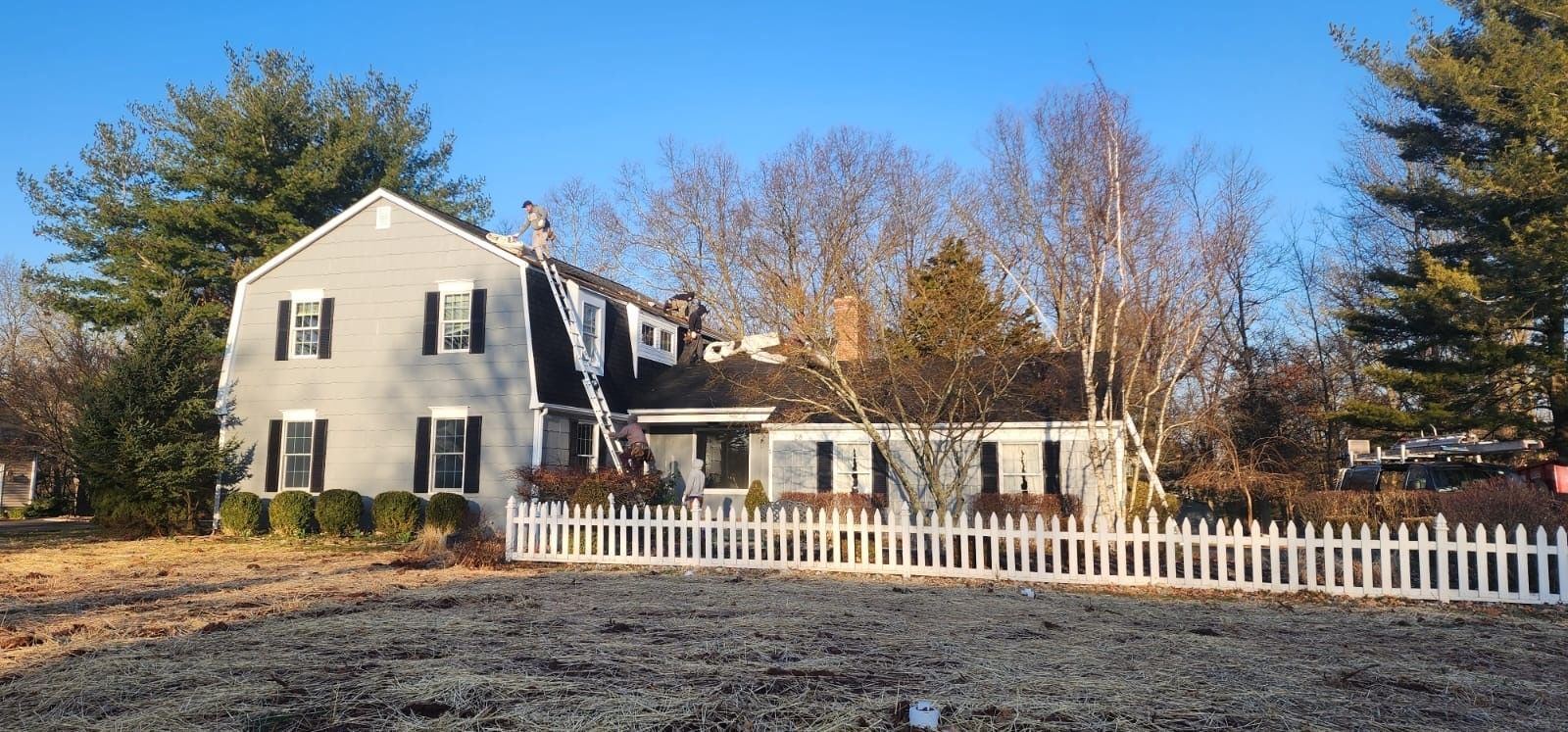 A house with a white picket fence, surrounded by trees under a blue sky; ladders on the roof.