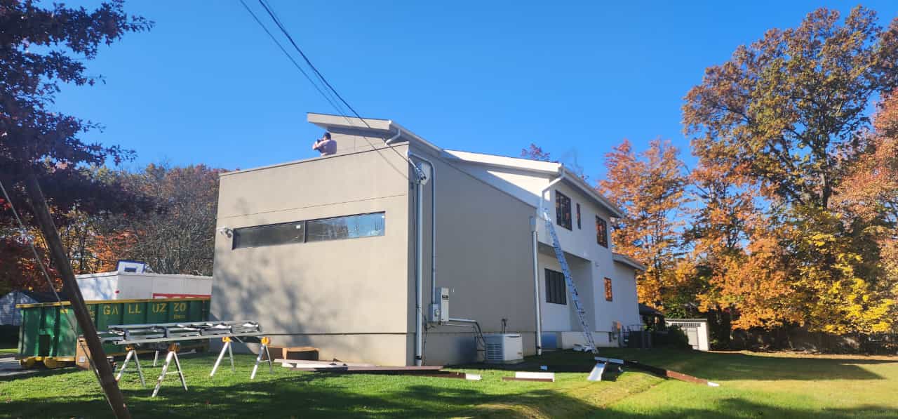 Two-story house with tan stucco and white siding, blue sky, and fall foliage. Construction materials are visible.