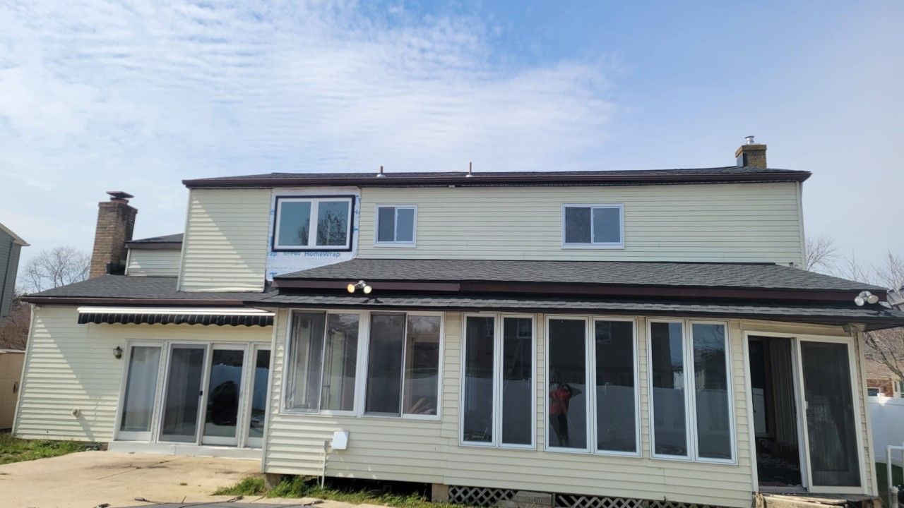 Back view of a two-story beige house with a sunroom, windows, and chimneys against a blue sky.