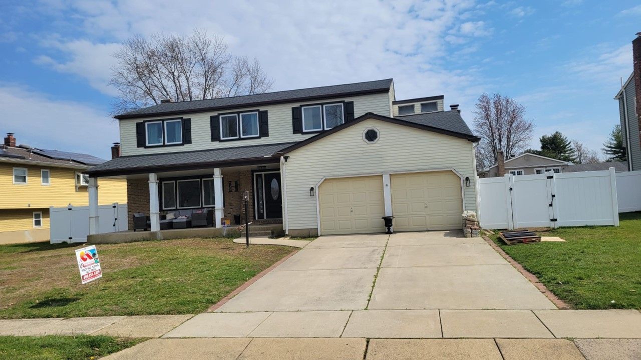 Two-story house with a driveway. Beige siding, black roof, and two-car garage. Green lawn and white fence.