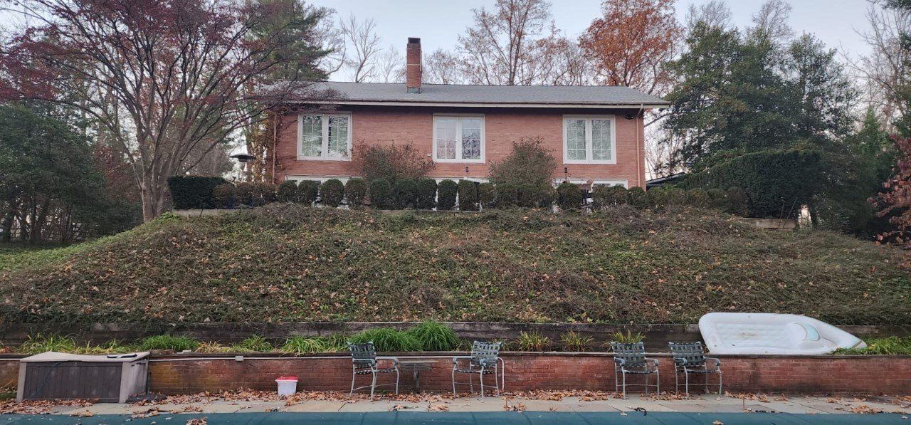 Brick house atop a grassy hill. Below, a pool with chairs. Overcast sky.