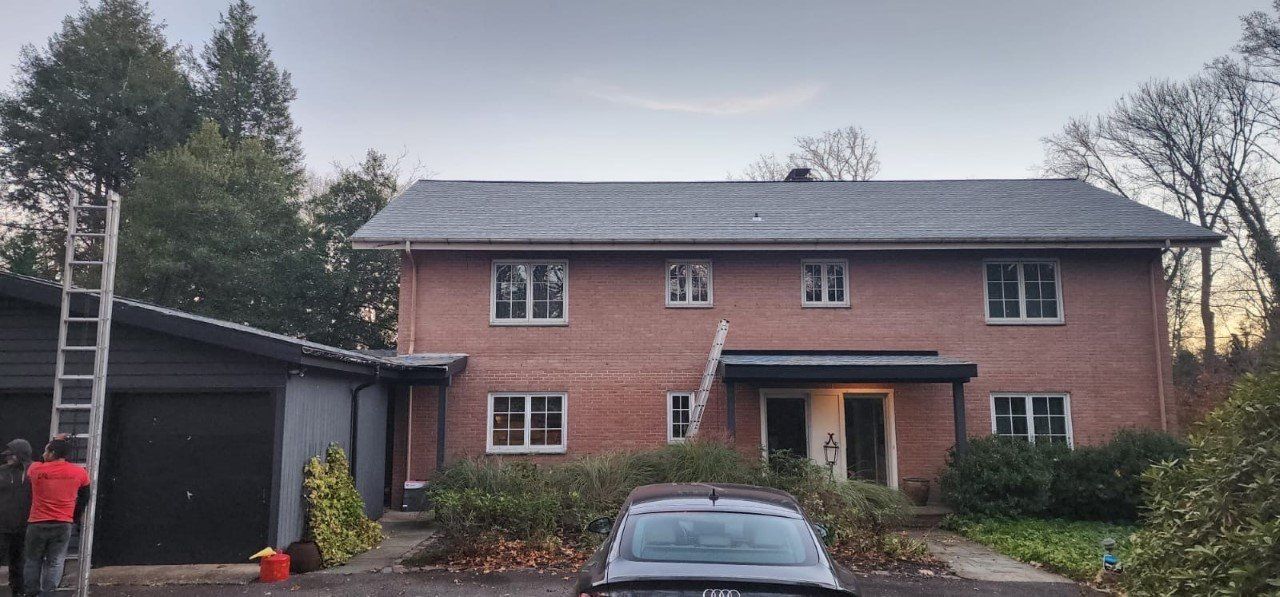 Two-story brick house with a dark gray roof. A ladder is leaning against the house, and a garage is to the left.