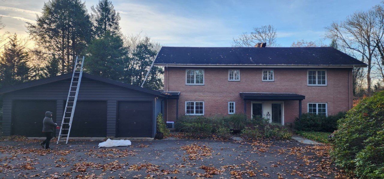A two-story brick house with a garage and a ladder. Leaves on the ground. A person is near the ladder.