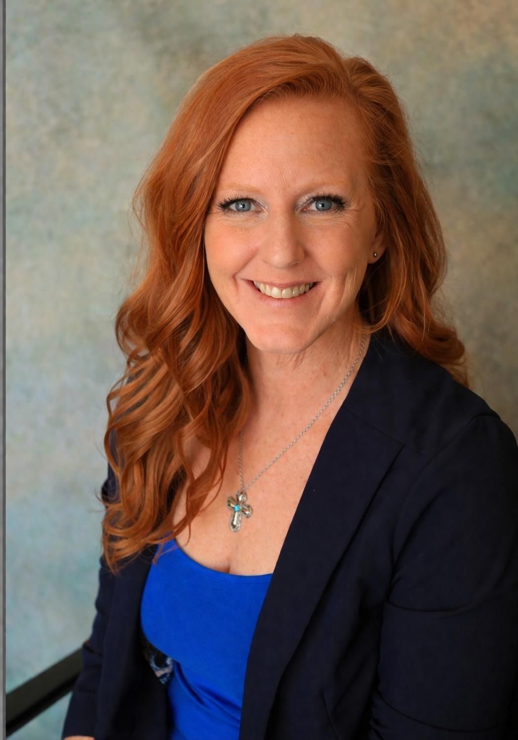 A woman with long red hair smiles while wearing a blue top, dark blazer, and cross necklace against a blue-grey backdrop.
