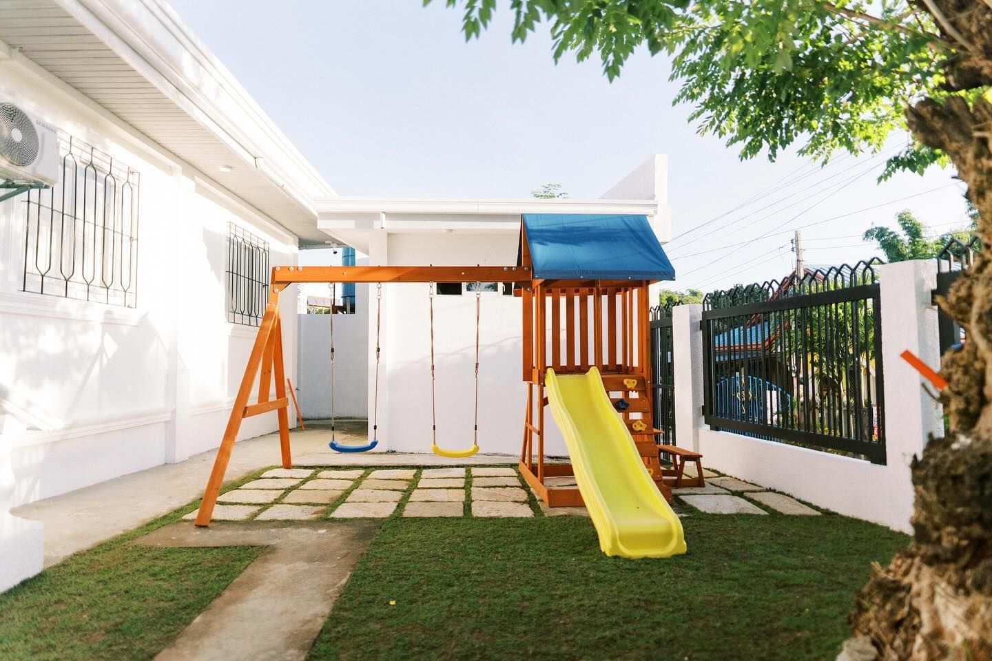 Playground with slide and swings in a grassy yard, next to a white building.