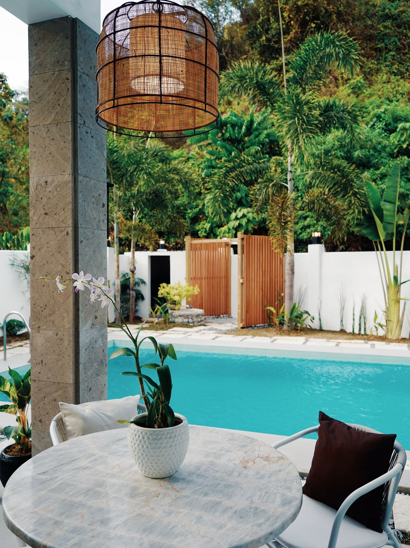 Outdoor patio with pool. Round table with white chairs, view of pool and lush greenery. Woven light fixture.